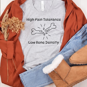 May include: A light gray t-shirt with the text "High Pain Tolerance, Low Bone Density" and a graphic of two broken bones. The shirt is paired with blue jeans, brown boots, and a rust-colored jacket.