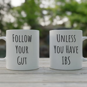 May include: Two white coffee mugs with black text. The mug on the left says "Follow your gut". The mug on the right says "Unless you have IBS".