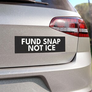 May include: A black rectangular bumper sticker with the white text "FUND SNAP NOT ICE" is affixed to the back of a silver car. The car's taillight is visible on the right side of the image.