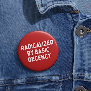 May include: A red button pin with the text "RADICALIZED BY BASIC DECENCY" in white lettering. The pin is set against a blue denim jacket, with visible stitching and a button. The pin is round and appears to be made of metal or plastic.
