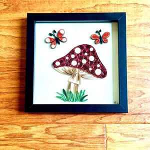 Puede incluir: Una obra de arte en quilling enmarcada que presenta una seta burdeos con lunares blancos y dos mariposas rojas y negras. La obra de arte está sobre un fondo blanco y enmarcada en un marco cuadrado negro. La obra de arte se exhibe sobre una superficie de madera.