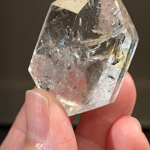 Puede incluir: Un cristal de cuarzo transparente con forma hexagonal. El cristal se sostiene en una mano y tiene inclusiones e imperfecciones internas. El cristal es transparente con un ligero tinte amarillo.