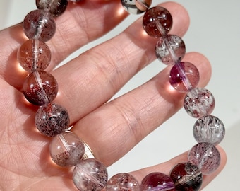 May include: A bracelet made of clear, round beads with internal inclusions of red, purple, and white. The beads are strung together to form a circular shape. The bracelet is held in a hand.