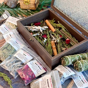 May include: A wooden box with a glass lid, filled with dried herbs, cinnamon sticks, and rose petals. Several small plastic bags containing herbs and labeled "Sage", "Rose Petals", and "Rosemary" are arranged around the box. Two bundles of sage are also visible.