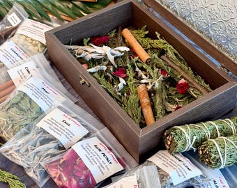 May include: A wooden box with a glass lid, filled with dried herbs, cinnamon sticks, and rose petals. Several small plastic bags containing herbs and labeled "Sage", "Rose Petals", and "Rosemary" are arranged around the box. Two bundles of sage are also visible.