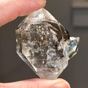 Op de afbeelding: Een helder, facetgeslepen kwartskristal met een rokerige tint. Het kristal heeft een complexe geometrische vorm met meerdere facetten en een kleinere kristalformatie. Het oppervlak reflecteert licht, waardoor een glinsterend effect ontstaat. Het kristal wordt vastgehouden door de vingers van een persoon.
