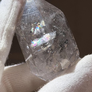 Puede incluir: Una punta de cristal de cuarzo transparente sostenida en un guante blanco. El cristal tiene una forma facetada con arcoíris e inclusiones internas. La luz se refracta a través del cristal, creando exhibiciones coloridas. El cristal es una gema natural y en bruto.