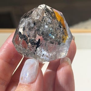Op de afbeelding: Een heldere kwartskristal met zwarte en gele insluitsels. Het kristal wordt in een hand gehouden en toont zijn veelzijdige structuur en natuurlijke schoonheid. De kwarts is een edelsteen.