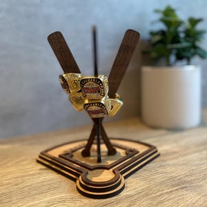 May include: A wooden baseball-themed display stand with two crossed bats and a baseball diamond base. The stand is holding four gold-colored rings with red "MVP" lettering.