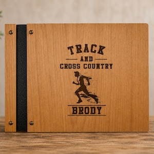May include: A rectangular wooden photo album with a dark brown leather spine secured by four metal rivets. The front cover features a laser-engraved graphic of a runner with the text TRACK AND CROSS COUNTRY and the name BRODY centered below.
