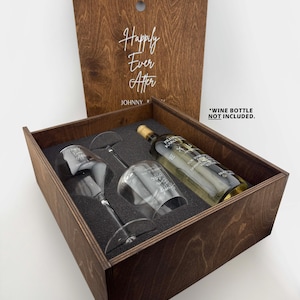 May include: A dark brown wooden gift box with a hinged lid featuring the text "Happily Ever After" and "JOHNNY". Inside, a dark foam insert holds two clear glass wine glasses and a wine bottle, each etched with text. Includes "WINE BOTTLE NOT INCLUDED."