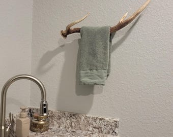 May include: A towel rack made from a set of antlers mounted on a white wall. A green towel is hanging on the rack. A stainless steel sink with a faucet is in the foreground.