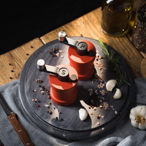May include: Two red salt and pepper grinders with black handles and silver tops. The grinders are on a dark grey round surface with pink and black peppercorns.