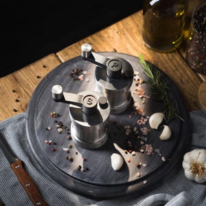 Puede incluir: Dos molinillos de pimienta y sal plateados con mangos negros y etiquetas en una tabla de madera gris oscuro con granos de pimienta rosa y negra, ramitas de romero y dientes de ajo.