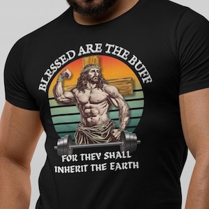 Pode incluir: Camiseta preta com um gráfico de estilo vintage de um Jesus musculoso levantando uma barra. O texto diz "Blessed are the Buff, for they shall inherit the earth."
