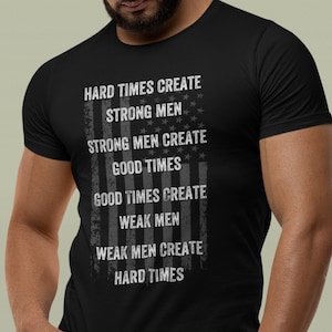 Può includere: Maglietta nera con un design di bandiera americana usurato e il testo "Hard times create strong men, strong men create good times, good times create weak men, weak men create hard times."