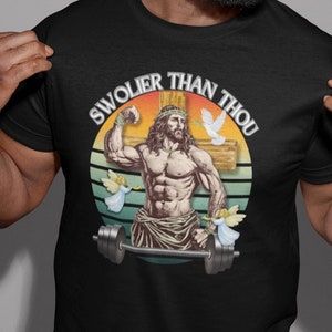 Pode incluir: Uma camiseta preta com um gráfico retrô de um Jesus musculoso levantando uma barra. O texto "Swolier Than Thou" está escrito em um arco circular ao redor da imagem.
