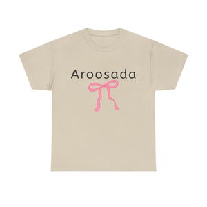 May include: A light tan t-shirt with the word "Aroosada" in black letters above a pink bow graphic. The t-shirt is made of a soft material and has a classic crew neck design.