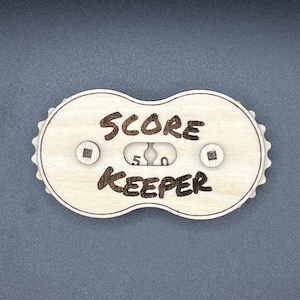 May include: A wooden score keeper with a counter that reads "5 0". The counter is surrounded by the words "SCORE KEEPER" in a handwritten font.
