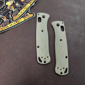 May include: Two green knife scales with gold screws. The scales are made of a textured material and have a rounded shape.