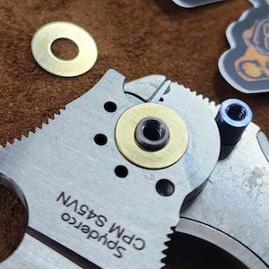 May include: A partially disassembled Spyderco CPM S45VN knife with a brass washer in place. Several additional brass washers are scattered nearby. Two stickers with the text "OUTLAW COMMAND POST" are also visible.