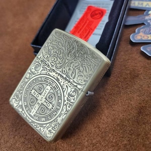 May include: A brass-colored lighter with an ornate engraved design, featuring a circular emblem with text and a cross. The lighter is in an open black box, with a red tag and a paper insert visible. The words "Made in USA" are printed on the box.