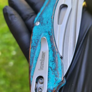 May include: A close-up of a folding knife with a weathered turquoise and brown handle. The blade is silver, and the pocket clip is marked "Kershaw". The knife is held against a blurred green background.