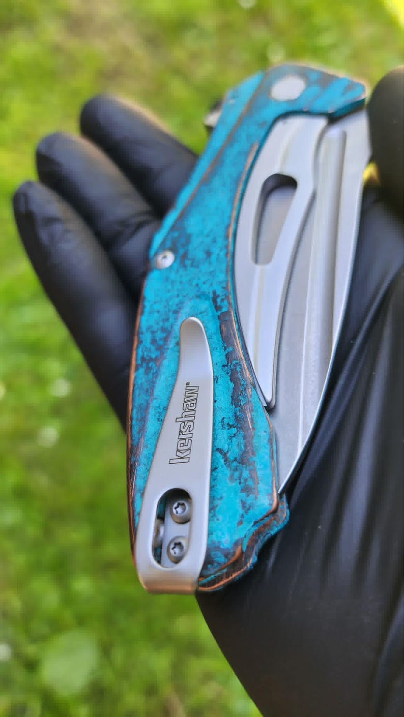 May include: A close-up of a folding knife with a weathered turquoise and brown handle. The blade is silver, and the pocket clip is marked "Kershaw". The knife is held against a blurred green background.
