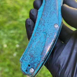 May include: A folding knife with a weathered, turquoise-colored handle and a silver blade. The handle has a textured, aged appearance with hints of brown. The blade is partially open, revealing its sharp edge. The knife is held against a blurred green background.