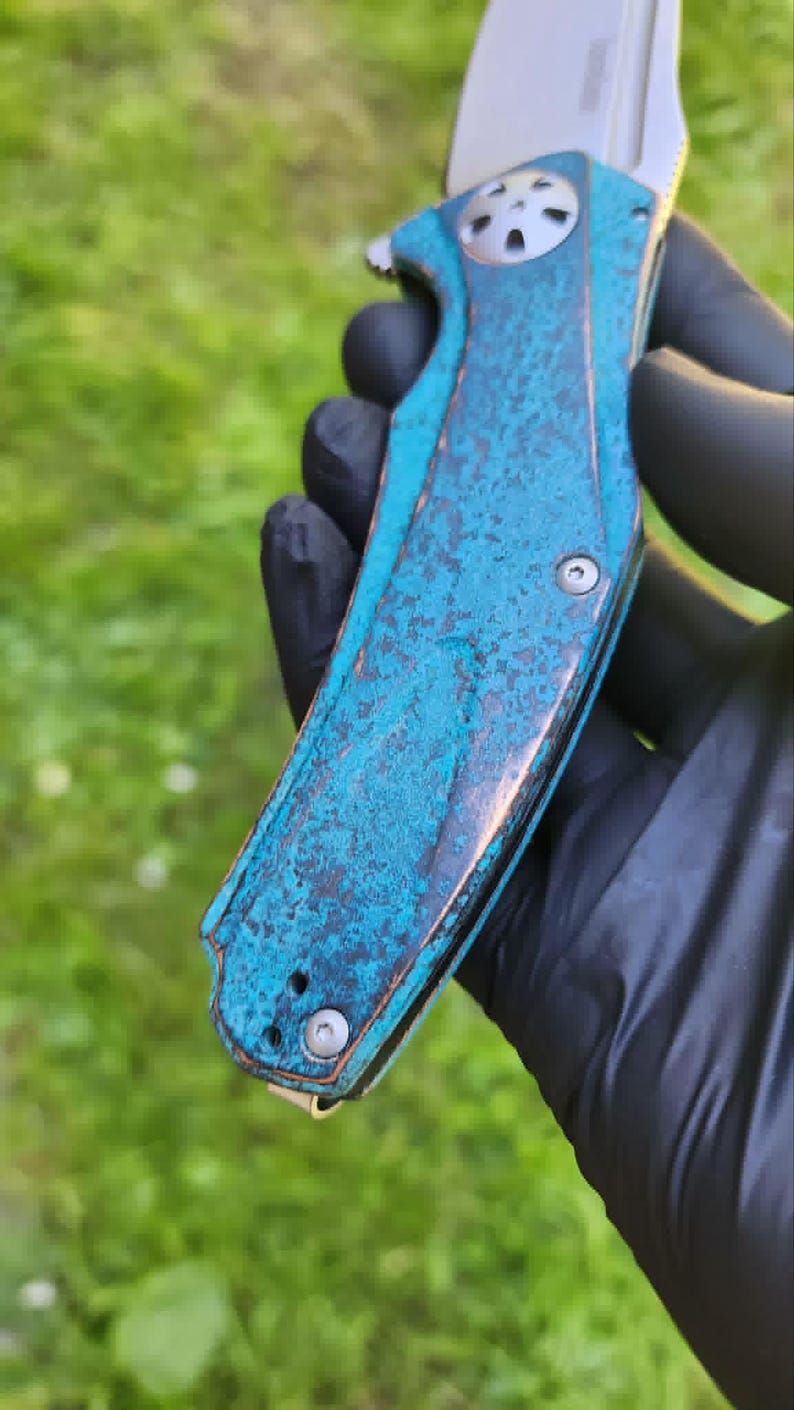 May include: A folding knife with a weathered, turquoise-colored handle and a silver blade. The handle has a textured, aged appearance with hints of brown. The blade is partially open, revealing its sharp edge. The knife is held against a blurred green background.