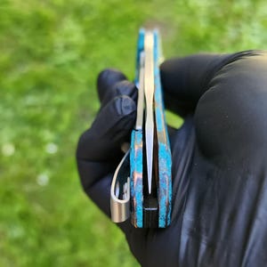 May include: A pocket knife with a blue and bronze handle and a silver blade. The knife is held in a gloved hand, with the blade partially open. The handle has a textured appearance.