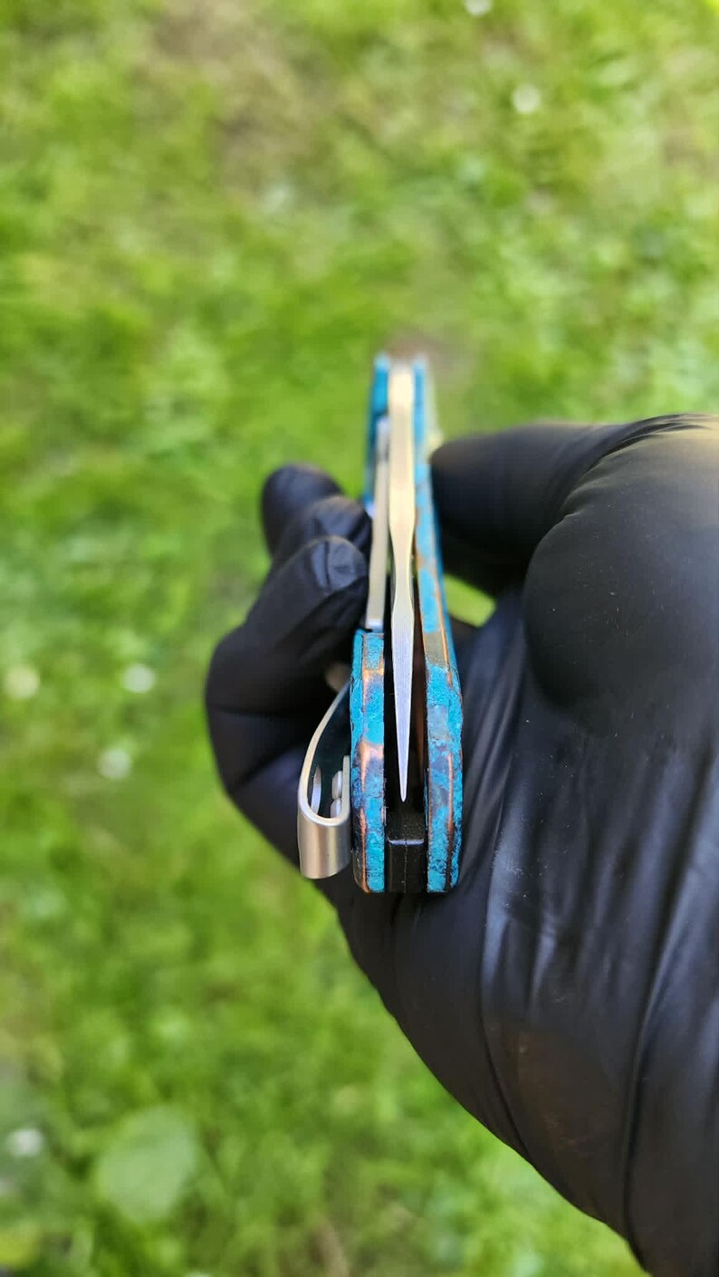 May include: A pocket knife with a blue and bronze handle and a silver blade. The knife is held in a gloved hand, with the blade partially open. The handle has a textured appearance.