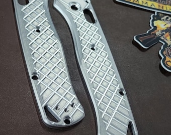 Könnte beinhalten: Zwei silberfarbene Messergriffschalen aus Metall mit einem texturierten rautenförmigen Gittermuster, vorgebohrten Schraublöchern und einem Lanyard-Loch an der Basis. Die Schalen werden von drei Aufklebern mit dem Text Outlaw Command begleitet.