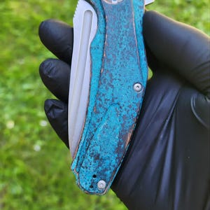 May include: A folding pocket knife with a weathered, turquoise-colored handle and a silver blade. The handle has a textured appearance with a circular design on the end. The knife is held in a black gloved hand against a green grassy background.