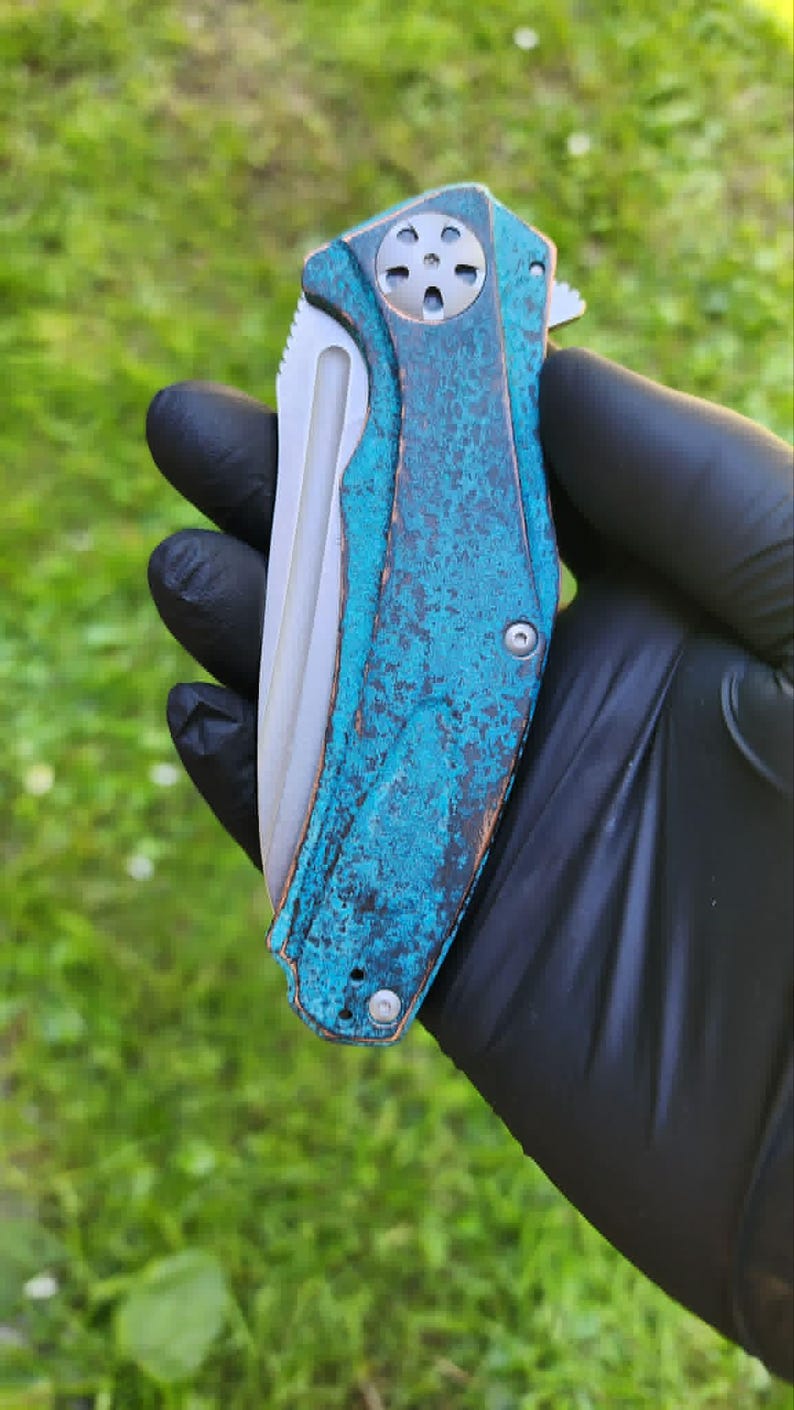 May include: A folding pocket knife with a weathered, turquoise-colored handle and a silver blade. The handle has a textured appearance with a circular design on the end. The knife is held in a black gloved hand against a green grassy background.