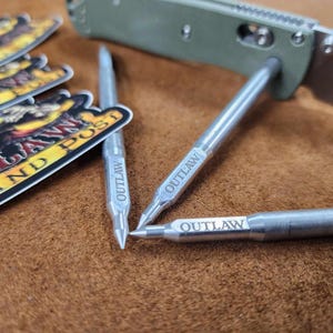 May include: Three silver-colored metal pens with pointed tips, each engraved with the word OUTLAW. One pen is positioned through the handle of a green folding knife. Three stickers with the text OUTLAW COMMAND POST are visible in the background.