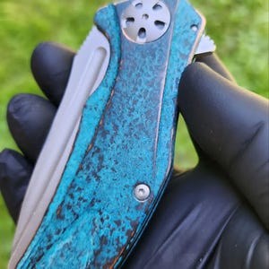 May include: A folding pocket knife with a weathered, turquoise and brown handle. The blade is silver, and the handle has a textured, aged appearance. The knife is held against a blurred green background.