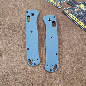May include: Two light blue knife handle scales with multiple holes and a textured surface. The scales are lying on a brown surface, with several "OUTLAW COMMAND POST" stickers in the background.