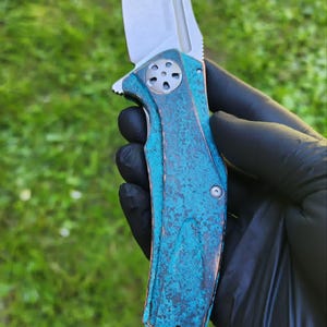 May include: A folding knife with a silver blade and a handle with a blue-green, textured finish. The knife is held against a green grassy background.