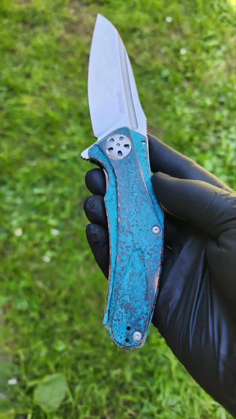 May include: A folding knife with a silver blade and a handle with a blue-green, textured finish. The knife is held against a green grassy background.