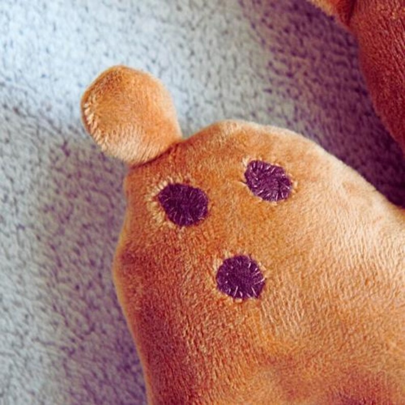 May include: Close-up of a plush toy's ear and head. The toy is a warm brown color with three dark purple spots and a small ear. The plush material has a soft, fuzzy texture.