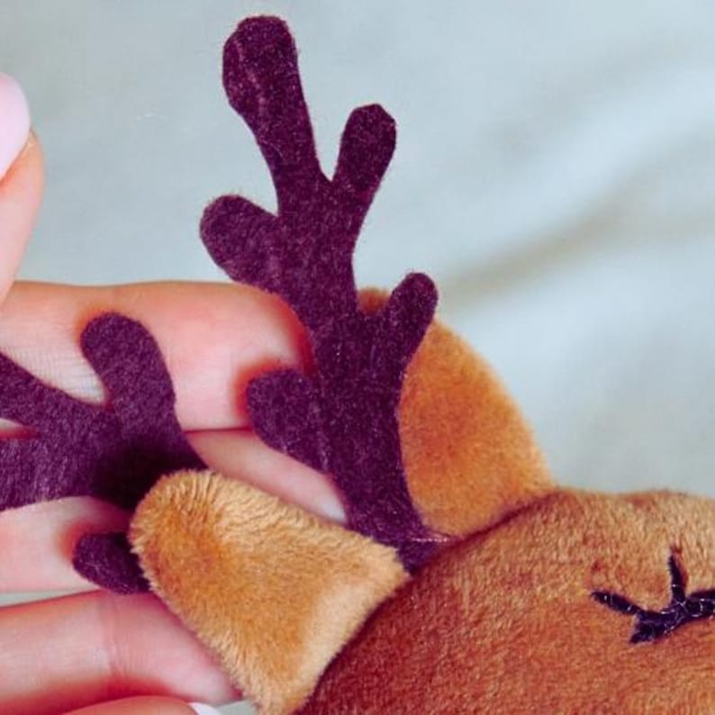 May include: Close-up of a plush reindeer head with brown fur and dark purple antlers. The reindeer has a light brown ear and a stitched black eye. The image is well-lit, highlighting the soft texture of the materials.