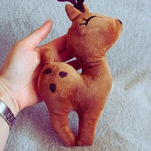 May include: A plush brown deer toy with black spots and antlers. The deer has closed eyes and a small tail. The toy is made of soft material and is held in a hand.