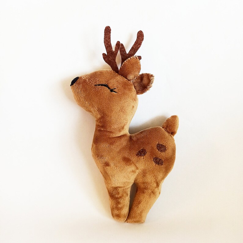 May include: A plush deer toy in shades of brown, with a soft, textured body. The deer has small, dark brown antlers, a small tail, and embroidered eyes. The toy features darker brown spots on its side.