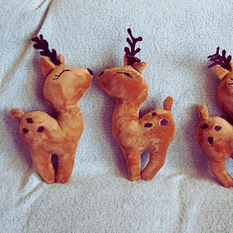 May include: Three plush reindeer toys in shades of brown with dark brown spots and antlers. The reindeer have closed eyes and are arranged on a light-colored surface, suggesting a festive or holiday theme.