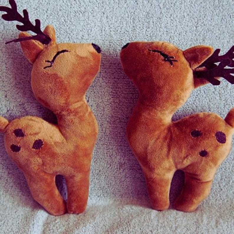 May include: Two plush deer toys in a warm brown color, each with dark brown antlers and black embroidered eyes. The deer have dark purple spots on their bodies and are positioned on a light blue surface.