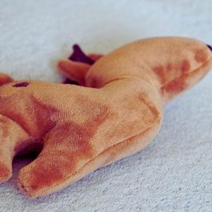 May include: A plush toy in the shape of a deer, lying on a light-colored surface. The deer is a soft, muted orange color with darker brown accents on the antlers and a small purple nose. The toy has a simple, rounded design.