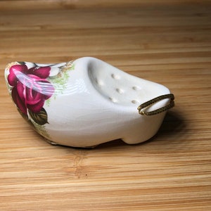 May include: White ceramic clog with a floral design featuring pink roses and green leaves. The clog has a gold loop handle and a series of holes on the top.
