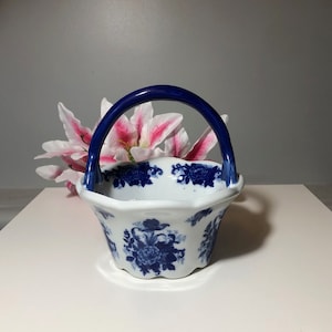 May include: A white ceramic basket with a blue floral design and a blue handle. The basket is sitting on a white surface.