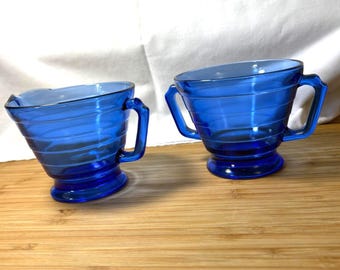 May include: A matching pair of cobalt blue glass cream and sugar vessels. Each piece has a handle and a spiral design. The sugar bowl has two handles, and the creamer has a spout. The set is on a wooden surface.